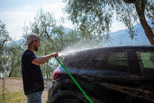 Man washes a black car with a hose at a sunny outdoor location surrounded by trees and mountains