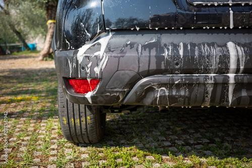 Car washing process happening in a garden area with soap suds on a vehicle rear and wet tire visible