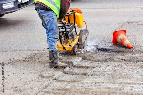 Worker operates machine to repair road while traffic cone stands nearby on busy street.