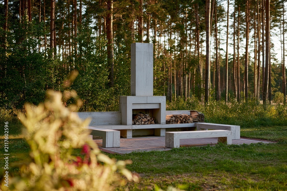 Fototapeta premium An outdoor fireplace seating area at the edge of pine forest, presented with stacked firewood under warm afternoon side light.