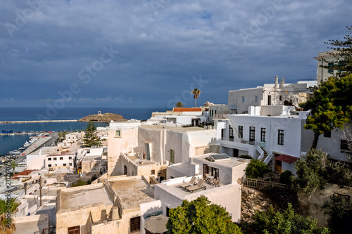 Elevated view across the whitewashed houses and harbor of Chora town, Naxos island, Greece, beneath dramatic storm clouds, with the aegean sea and coastal waterfront beyond.
