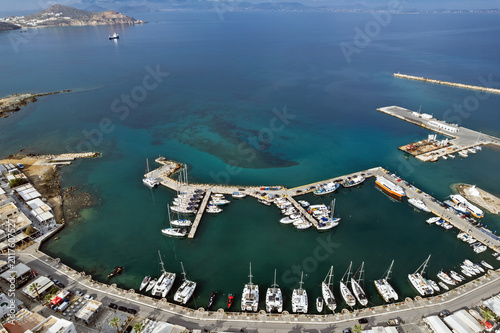 Aerial view of Chora harbor in Naxos island, Greece, with yachts, ferries and sailboats moored around curved piers in clear turquoise Aegean waters beneath distant cycladic islands.