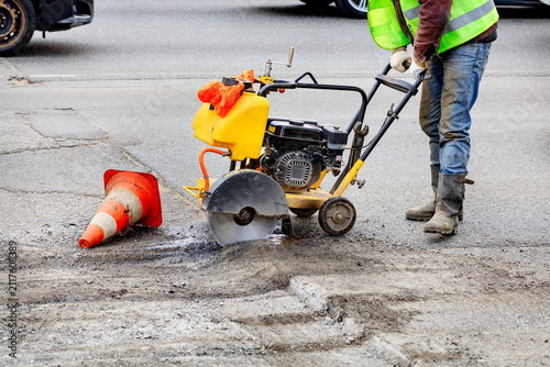 Worker cuts asphalt on busy road with concrete saw and traffic cones nearby in morning light.