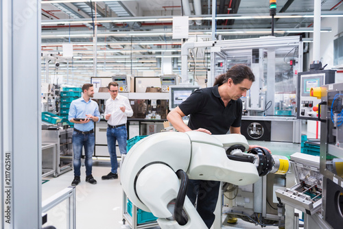 Man operating assembly robot in factory with two men in background supervising