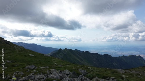 Aerial drone flight rising from a mountain summit toward dramatic clouds above the Făgăraș Mountains, with sunlight illuminating the upper clouds while their darker bases contrast against the distant 