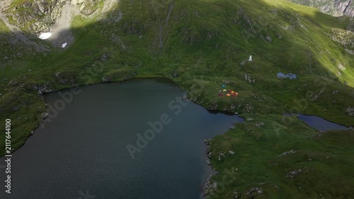 Forward descending drone flight toward a mountain lake surrounded by alpine peaks, with tents and visitors gathered near the water and a small patch of lingering snow visible in the landscape.