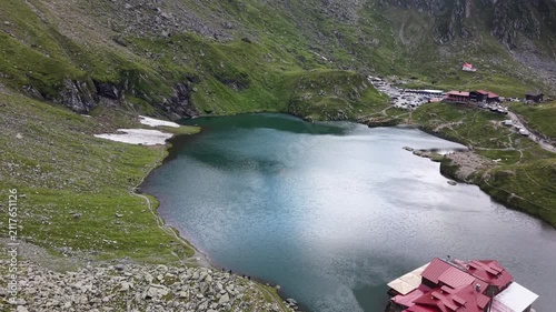 Drone flying forward above the famous Bâlea Lake mountain restaurant toward the parking area, capturing tourists, vehicles, remaining snow patches and spectacular alpine scenery in Romania's Mountains