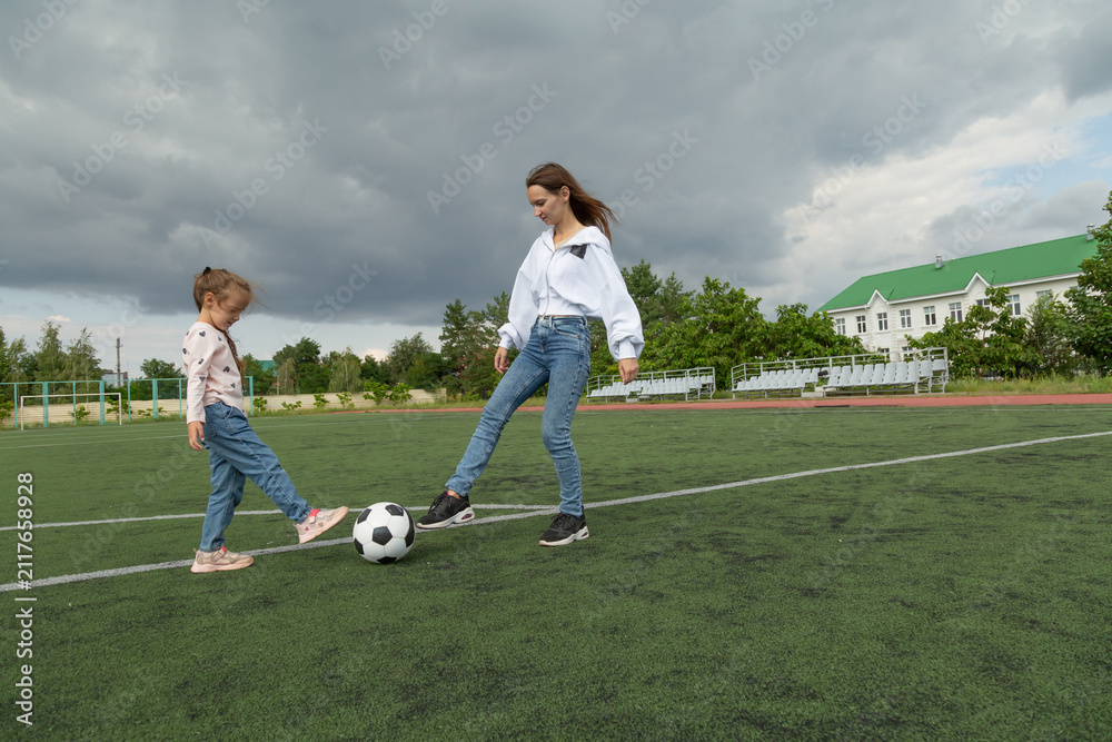 Obraz premium Young mother teaches girl soccer on green field, capturing motherhood in motion. Motherhood encourages learning, motherhood reflects care, and motherhood elevates sport storytelling campaigns.