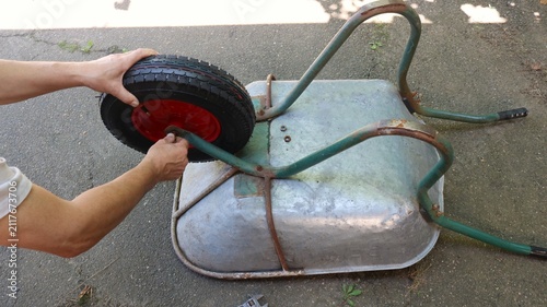 The process of repairing an overturned garden wheelbarrow on asphalt. A man installs a new inflatable tire with a black tread and a red disc onto the metal axle of the frame, securing it with his hand