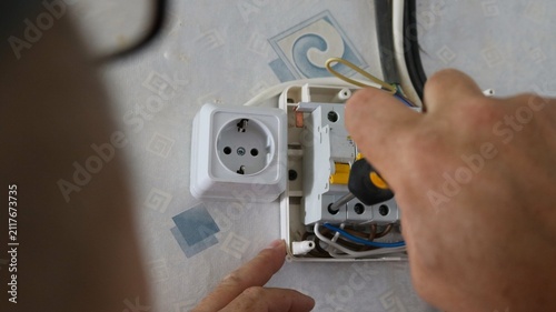 A close-up of an electrical panel installation. A technician's hand tightens a screw on a machine with a screwdriver. A white outlet is mounted nearby, seen from behind a blurred silhouette of a man