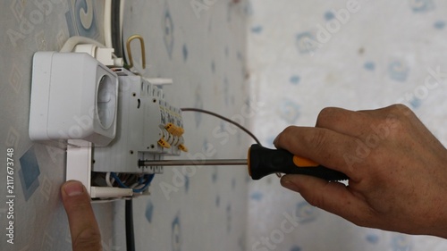 Installing a surface-mounted electrical panel on a wallpapered wall. A technician uses a screwdriver to tighten the contact on a circuit breaker. A white outlet is installed on the left.
