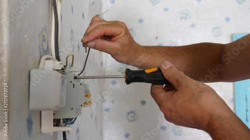 The hands of an electrician repairing electrical wiring. With one hand, the person holds a bare copper wire, while with the other, using a screwdriver, they install or test a circuit breaker.