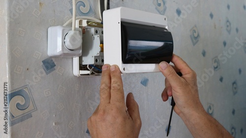 The process of installing an electrical panel on a wallpapered wall. Hands position the white plastic housing with a dark protective cover over the circuit breakers, holding a screwdriver nearby.