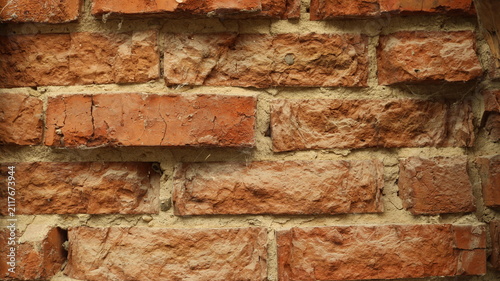 A close-up of an old red brick wall. The textured brickwork, aged and pitted, has uneven surfaces, chips, and small cracks, held together by rough cement mortar. Macro texture of damaged brickwork.
