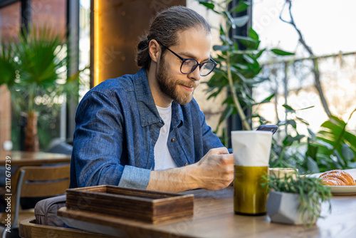 Fotografie Young bearded man in eyeglasses sitting at the table in a cafe