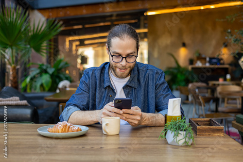 Foto Young bearded man in eyeglasses sitting at the table in a cafe