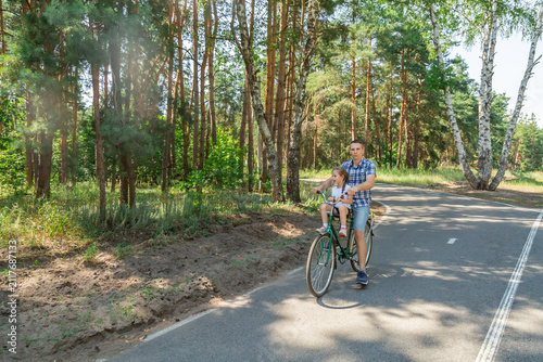 A young father carries his little daughter in a bicycle seat. Dad and child ride a bike in forest