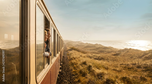 Travel train coastal landscape sunset ocean passenger window journey railway, scenic coastal train travel sunset with passenger leaning from window along railway beside ocean and dune landscape