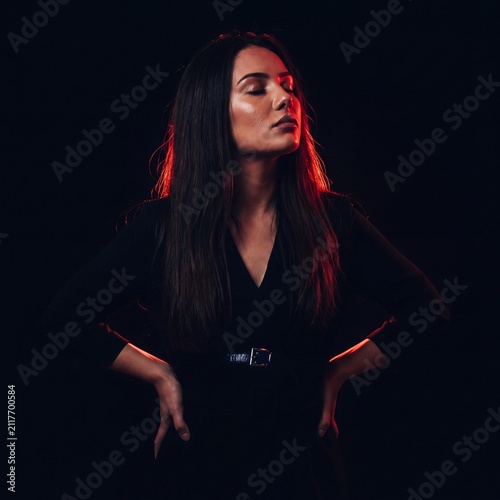 A beautiful young woman with long dark hair poses confidently against a black background, highlighted by dramatic red rim lighting.