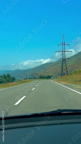 POV Car Driving on Scenic Highway Road Surrounded by Green Mountain Hills, Power Lines, and Blue Sky with White Clouds on a Sunny Summer Day