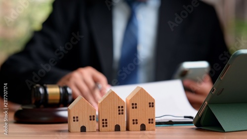 Quadro su tela Three small wooden house models sit near auction gavel refocused male attorney reviews real estate contract document holding smartphone device inside courtroom office