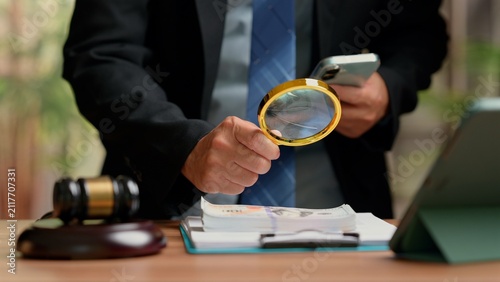 lawyer forensic investigator wearing suit holding magnifying glass examines dollar banknotes near judge gavel tablet lawyer office desk conducting financial fraud audit corruption law investigation.