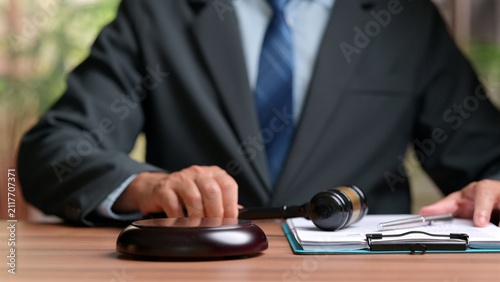 Male lawyer wearing formal suit holds wooden gavel near legal document clipboard paper sitting inside courtroom office workspace, representing justice system, law, trial, authority.