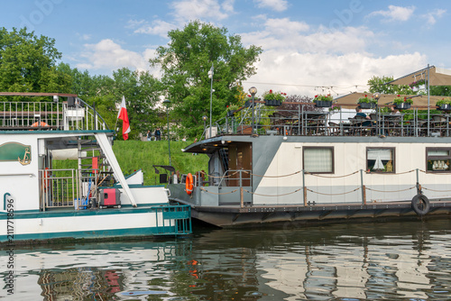 Принти на полотні Restaurant boats moored on the Vistula River in Krakow, Poland, feature an open-air dining terrace, flower boxes, sun canopies and a Polish flag