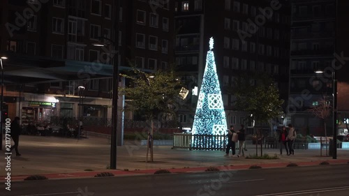 People walking by a flashing christmas tree in a city at night