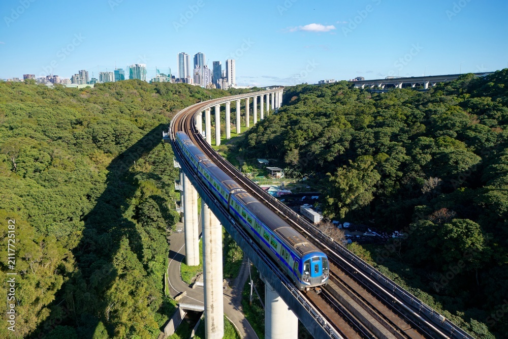 Fototapeta premium A metro train of Taoyuan Airport MRT travels on the elevated rail, crossing green wooded hills on a beautiful sunny day with high rise buildings in Linkou Dist. in backgroud in New Taipei City, Taiwan