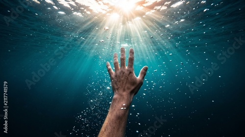 Hand reaching up towards sunlight rays from underwater, drowning person asking for help, hope salvation despair.