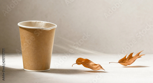Disposable paper cup with deformed rim and dried autumn leaves in soft breeze, bright studio