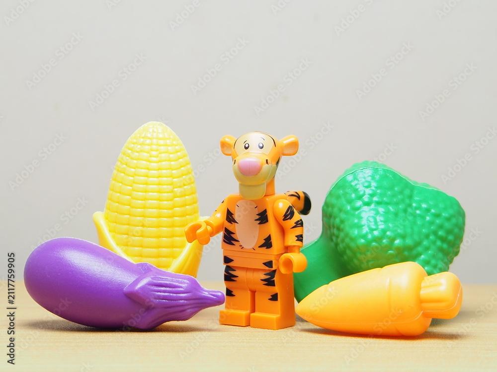 Fototapeta premium Indonesia - February 10, 2026 : Mini toy of action figure at outdoor with blur background. Miniature photography concept design. Minifigure of tiger guy character with eggplant, corn, carrot, broccoli