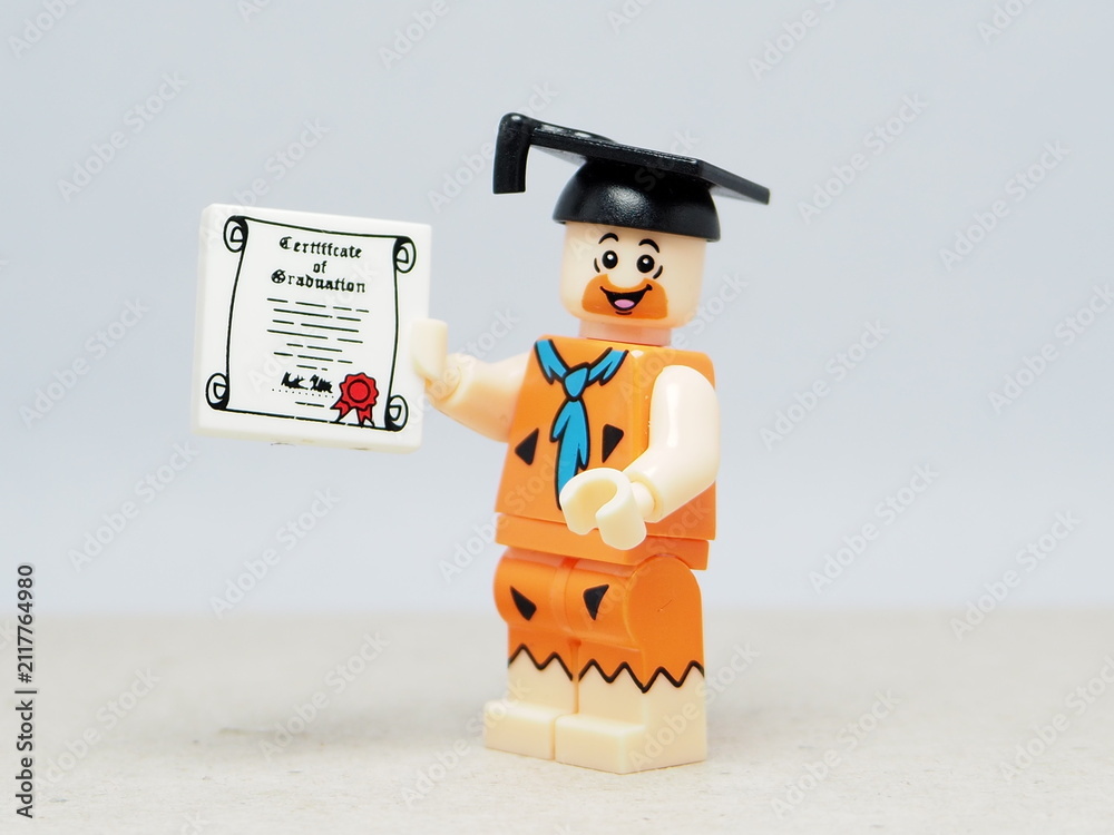 Obraz premium Indonesia - November 24, 2025 : Mini toy of action figure at table with blurred background. Miniature photography concept design. Minifigure of Fred Flintstone with graduation hat and certificate.