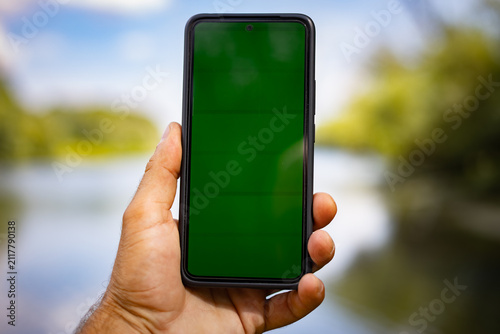 Close-up view of male hand holding smartphone with blank green screen against bokeh river and nature background for chroma key compositing and app presentation