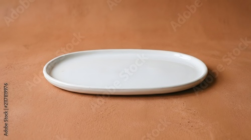 Empty white ceramic oval plate sitting centered on a textured warm terracotta surface featuring soft shadows and minimalist kitchen aesthetic