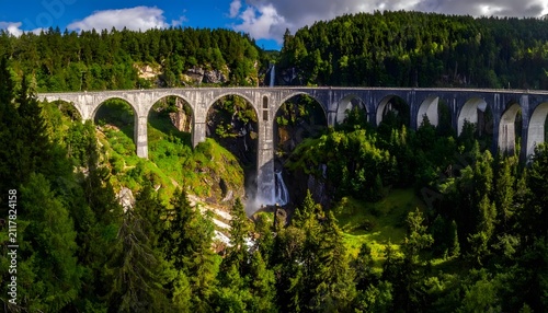 Stone railway viaduct stands tall over a lush green pine forest valley under a bright blue sky.