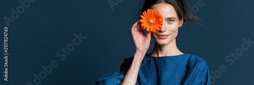 Young woman holding orange gerbera flower to eye in blue dress on dark background for fashion banner and editorial design