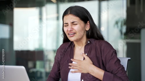 Young businesswoman suffering from intensive chest pain while working on laptop in modern office. Female employee holds her chest, experiencing discomfort, breathing difficulties, heart attack. Close 
