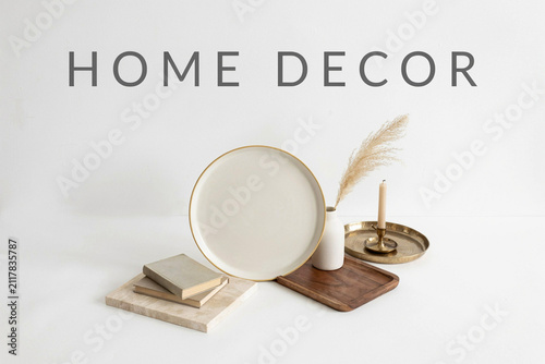 An elegant arrangement of various home decor items including a large cream plate a small white vase with pampas grass a lit candle and a stack of books creating a stylish interior display