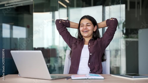 Happy young businesswoman finishes work on laptop while sitting at desk in modern business office. Relaxed female manager leans back in chair, feeling satisfied after completing daily tasks, projects.