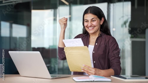 Happy young businesswoman receives good news approval letter while sitting in modern office. Smiling female employee is satisfied with positive notification, celebrates success, exciting achievement.