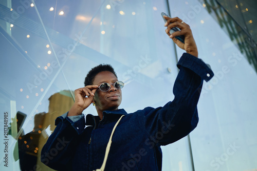Low angle shot of Black young adult woman holding smartphone and adjusting sunglasses while taking selfie outdoors, conveying mobile lifestyle and personal branding for social media marketing