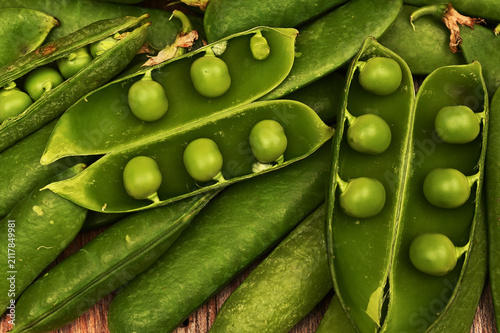 A close-up of fresh green pea pods, some opened to reveal ripe peas, arranged on a rustic wooden surface