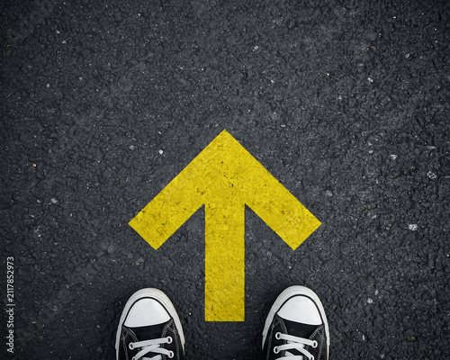 First person view of feet in sneakers standing before a painted yellow arrow on black asphalt pointing forward direction with direction pavement sneakers decision navigation opportunity