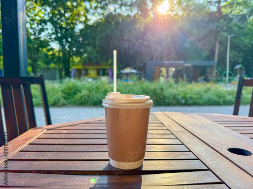 A warm beverage in a disposable cup sits on a wooden table, surrounded by green trees and sunlight in a park setting.