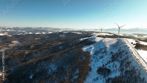 Aerial view of wind turbines on a snow-covered mountain range under a clear blue sky. Drone footage