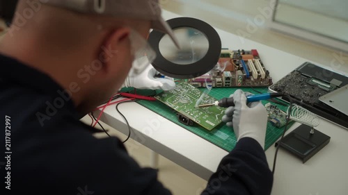 Electronics technician soldering a printed circuit board during electronic equipment repair. Precision engineering, PCB diagnostics, right to repair. Technology and electronics service.

