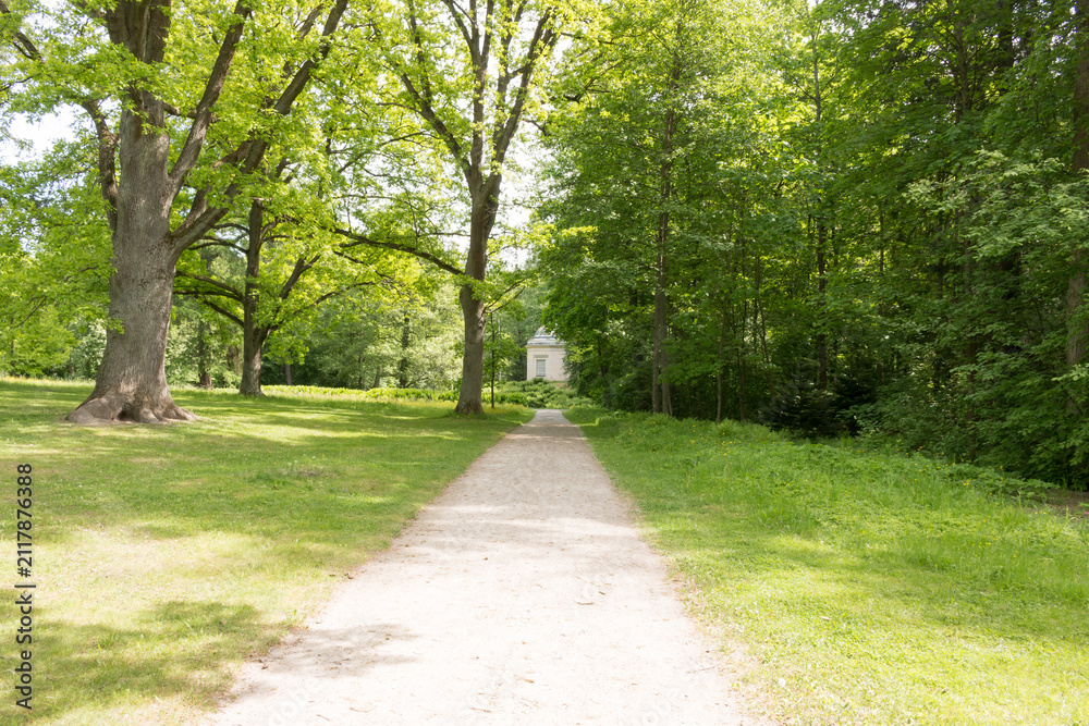 Obraz premium A scenic landscape view of historic Herttoniemi Manor Park in Helsinki, Finland, featuring sun-drenched paths, majestic old deciduous trees, and lush green lawns under a bright summer sky.