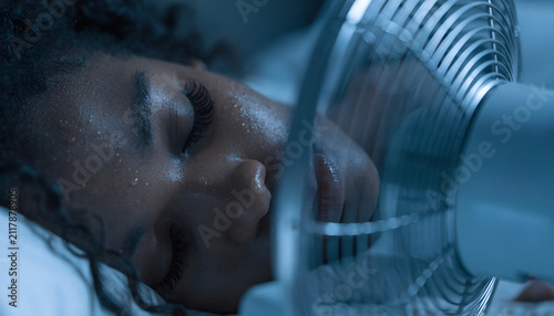 Young Black woman sleeping peacefully in a comfortable bed during a hot summer night, refreshed by the gentle breeze of a modern pedestal fan in a cozy dark bedroom.